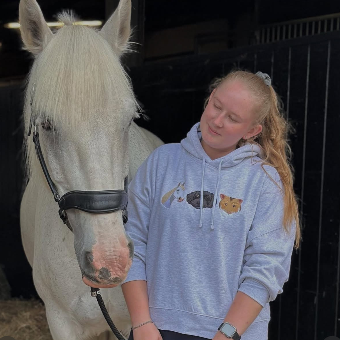 Girl in a blue hoodie with animal designs standing next to a white horse in a stable.