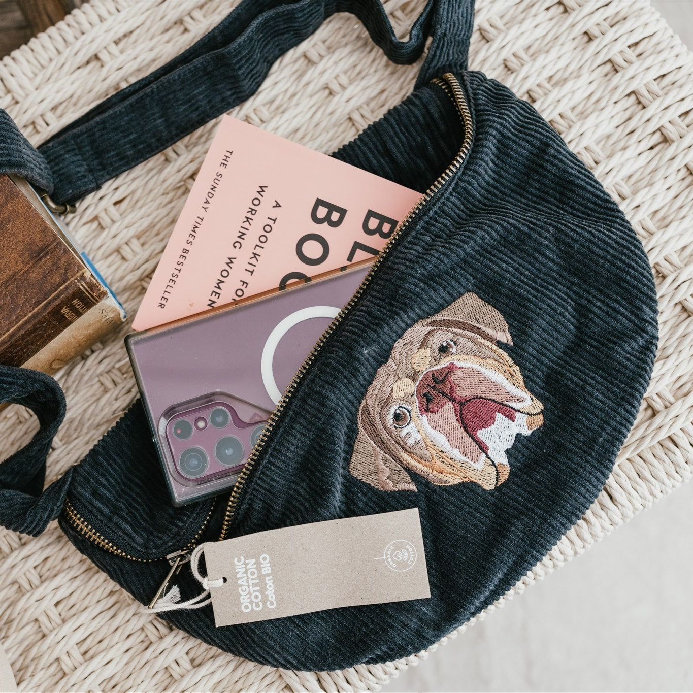 Black waist bag with bulldog design on a textured surface with books and phone.
